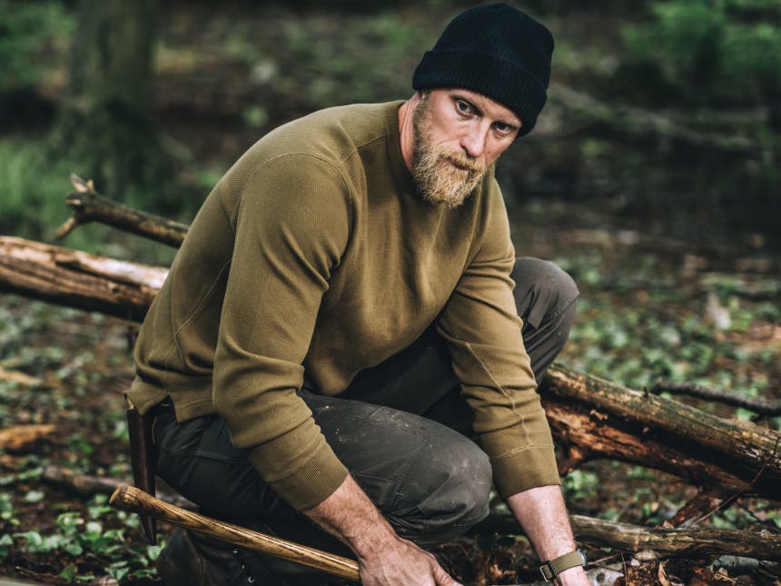 man in olive sweater, and black beanie, holding an axe kneeling on the forest floor