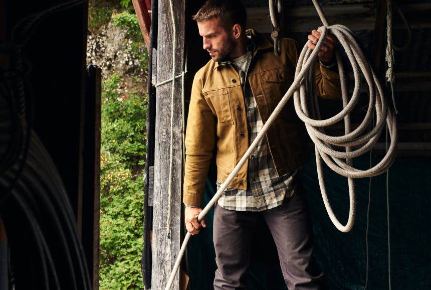man with tan waxed cotton coat and white and green shirt coiling large gauge rope in a barn