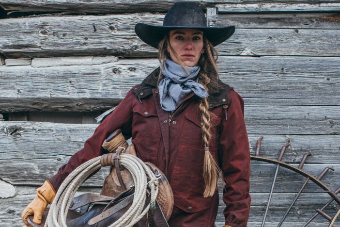 woman with gray cowboy hat, red coat, holds a saddle with a coil of rope while standing in front of a wooden wall