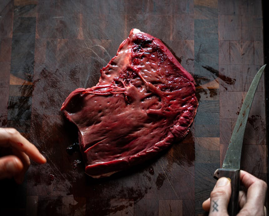 elk heart next to hand holding paring knife, fileted on a wooden cutting board