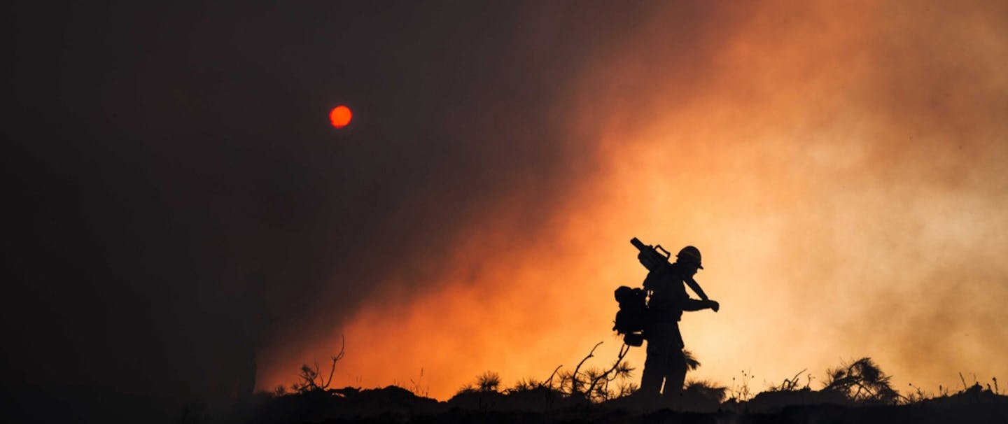 silhouette of wildland firefighter with large pack and gear slung over shoulder walking along ridgeline with smoke and red sun in background