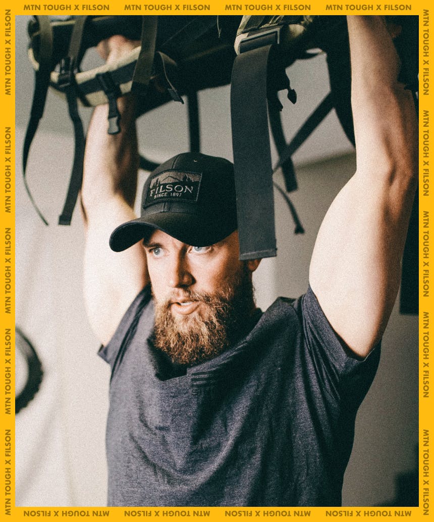 man lifting a backpack above his head in a black filson hat