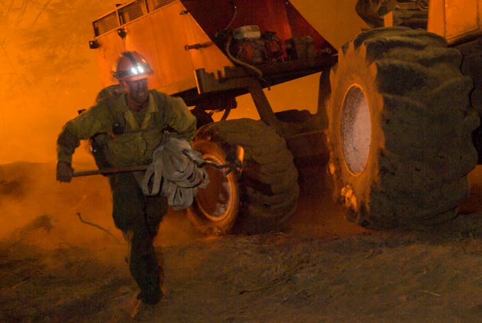wildland firefighter running holding a bundle of material in front of some large machinery reflecting the orange glow of the fire