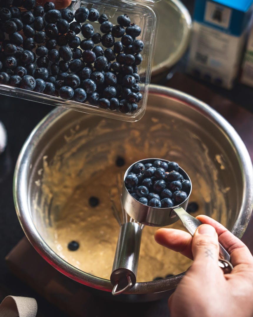 hand holding a measuring cup full of blueberries over a stainless steel bowl of batter