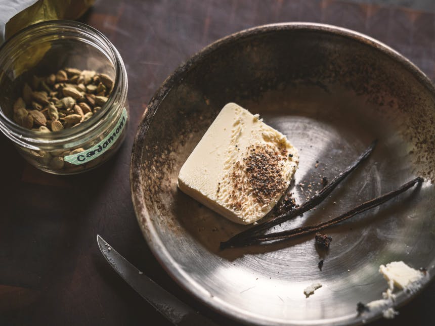 butter dusted with spices, split vanilla bean in a stainless steel dish, whole cardamom pods in a jar on the side on a wood cutting board