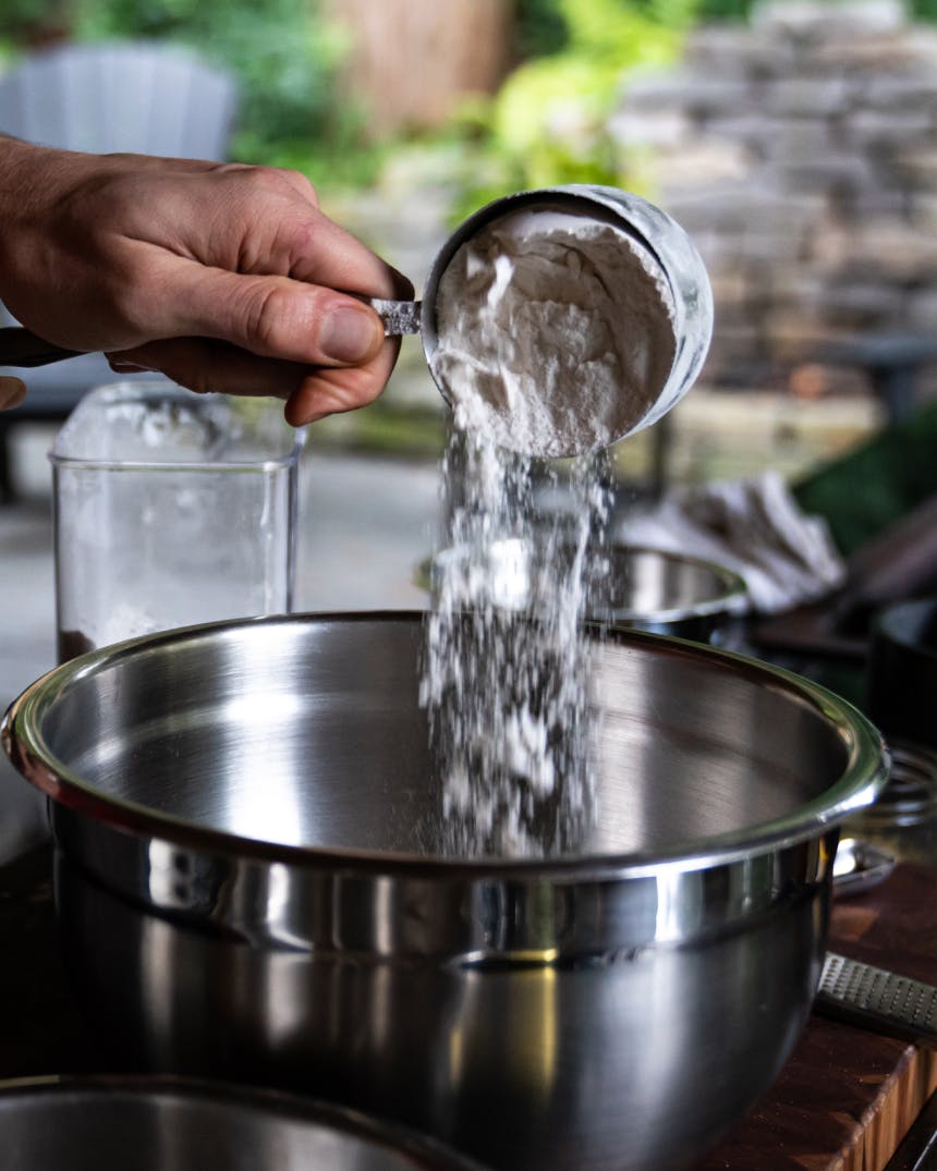 hand holding a measuring cup pouring flour into a stainless steel bowl