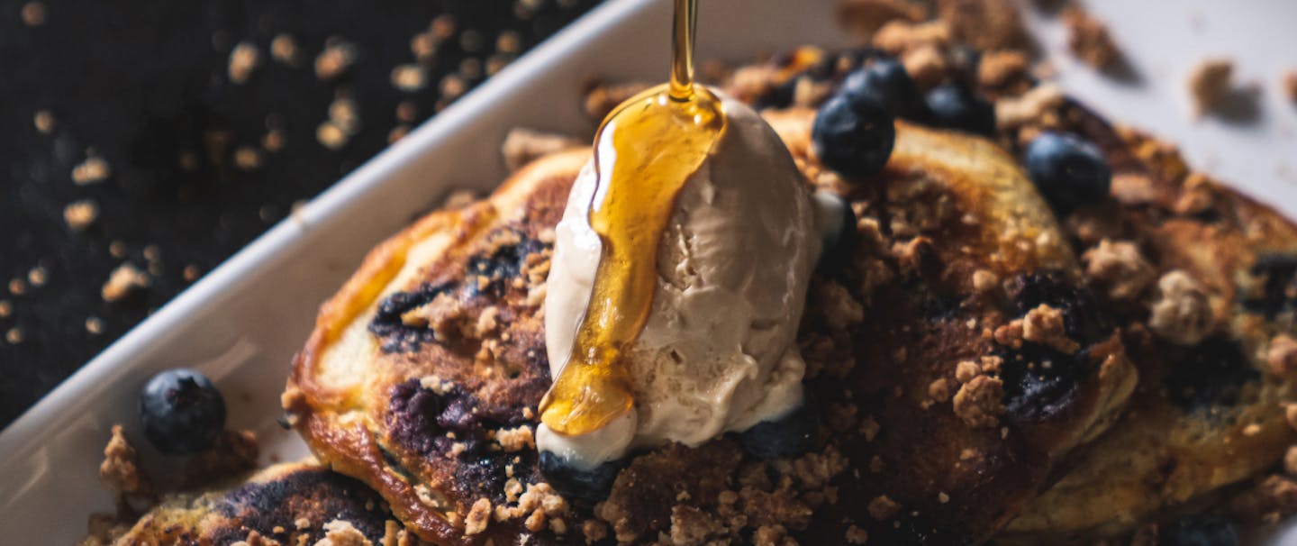 blueberry pancakes with crumbled walnuts on a white plate with syrup drizzling from a tin