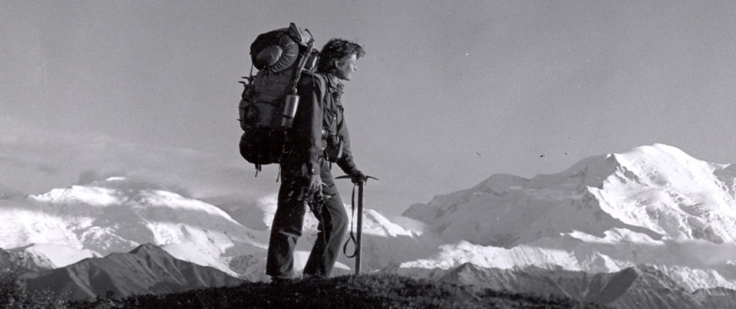 black and white image of person with pick and large mountaineering pack standing on a rocky peak with large snowy mountains in background