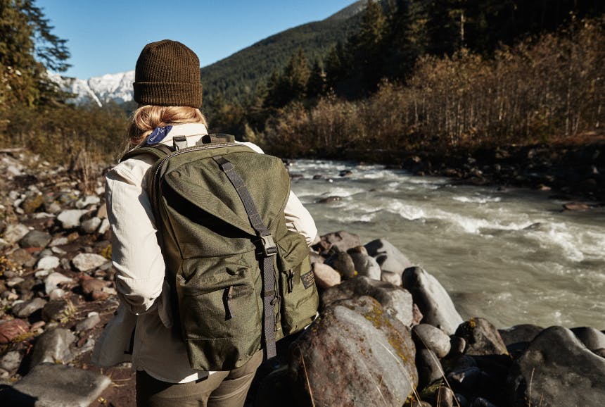 woman with brown beanie wearing green filson nylon ripstop backpack walking along the rock lined bank of a river