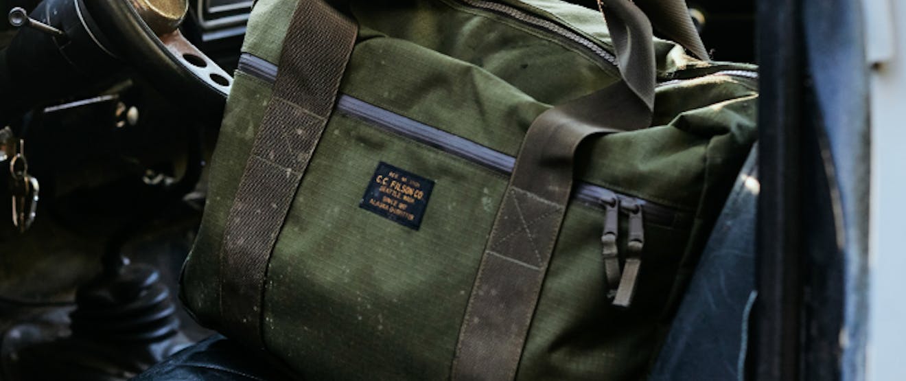 green filson duffel with olive straps, sits on a black leather car seat behind a metal and black plastic steering wheel