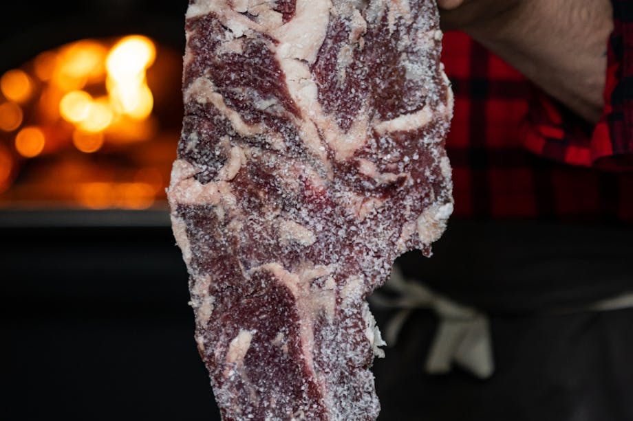 hand holding a cut of venison bacon uncut covered in a white powder in front of a wood fired stove