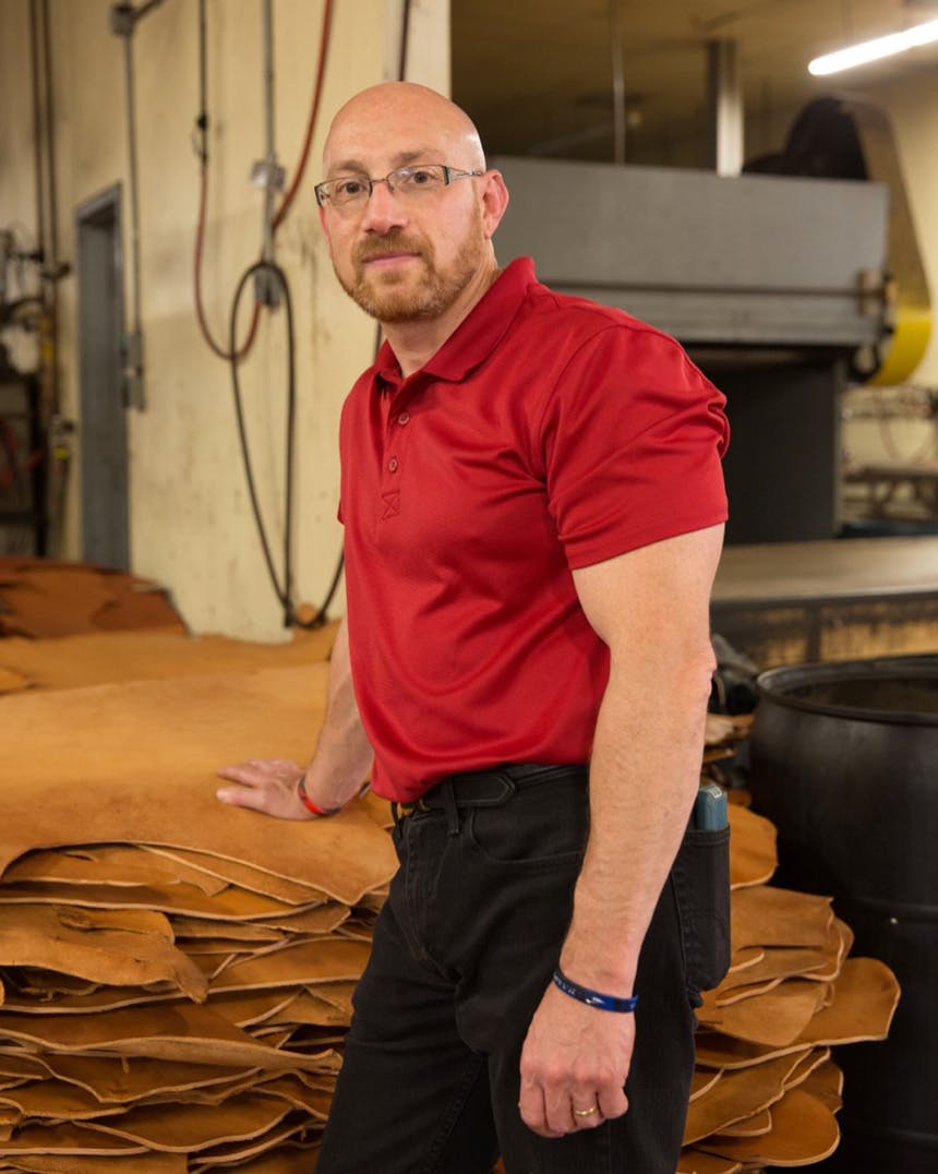 man with red polo shirt and black pants stands with hand on large stack of pieces of leather