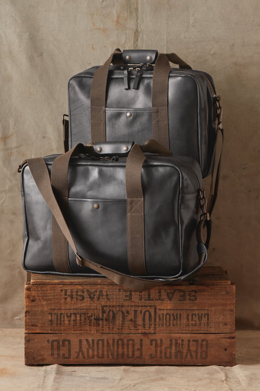 wickett and craig x filson black leather bags with olive straps sitting on top of wooden box