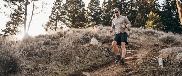 man in gray sweatshirt and black shorts running down a rocky hill in a high desert conifer forest