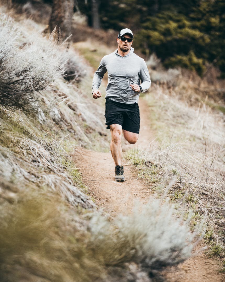 man in gray sweatshirt and black shorts running down a dirt trail on a hillside