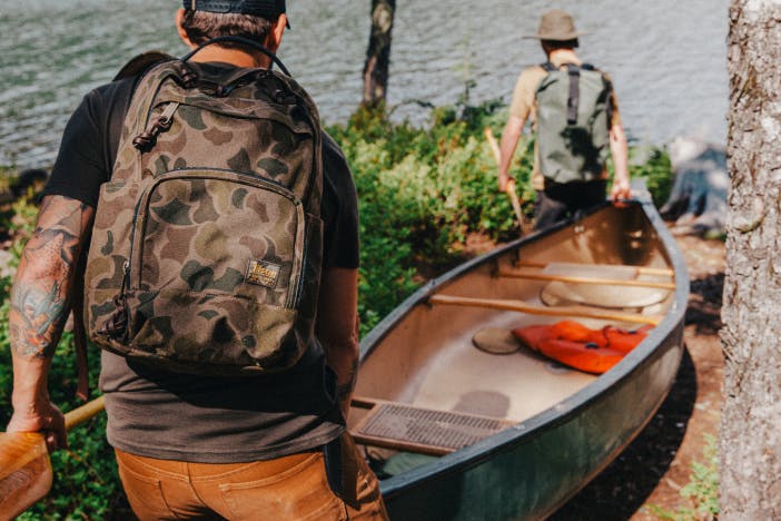 two men with filson backpackpacks on carry a green canoe down a dirt trail toward a river