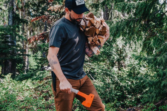 man holding a a small orange hatchet and an arm full of wood in a pine forest