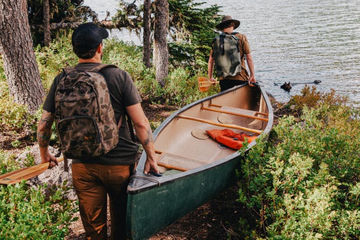 two men with filson backpackpacks on carry a green canoe down a dirt trail toward a river