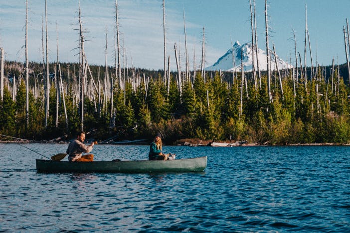two people paddling on a green canoe in a lake in a forest with old dead trees and new low lying pine growth with a snow covered mountain peak in the distance