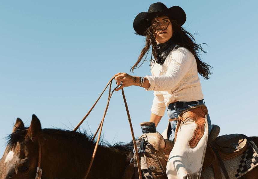 woman with black cowboy hat and white chaps and white shirt riding horse