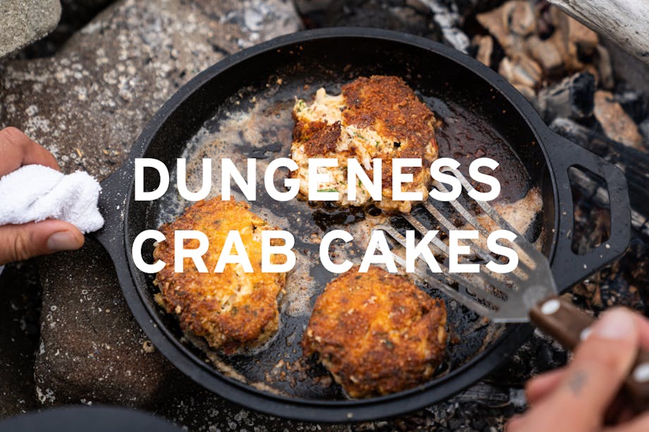 hand holding a cast iron skillet with three dungeness crab cakes over the coals of a fire central text reads 
