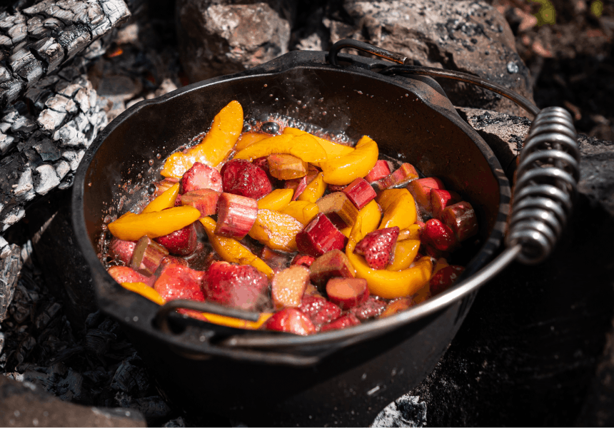dutch oven sitting on top of coals with peach and rhubarb and strawberries cooking inside