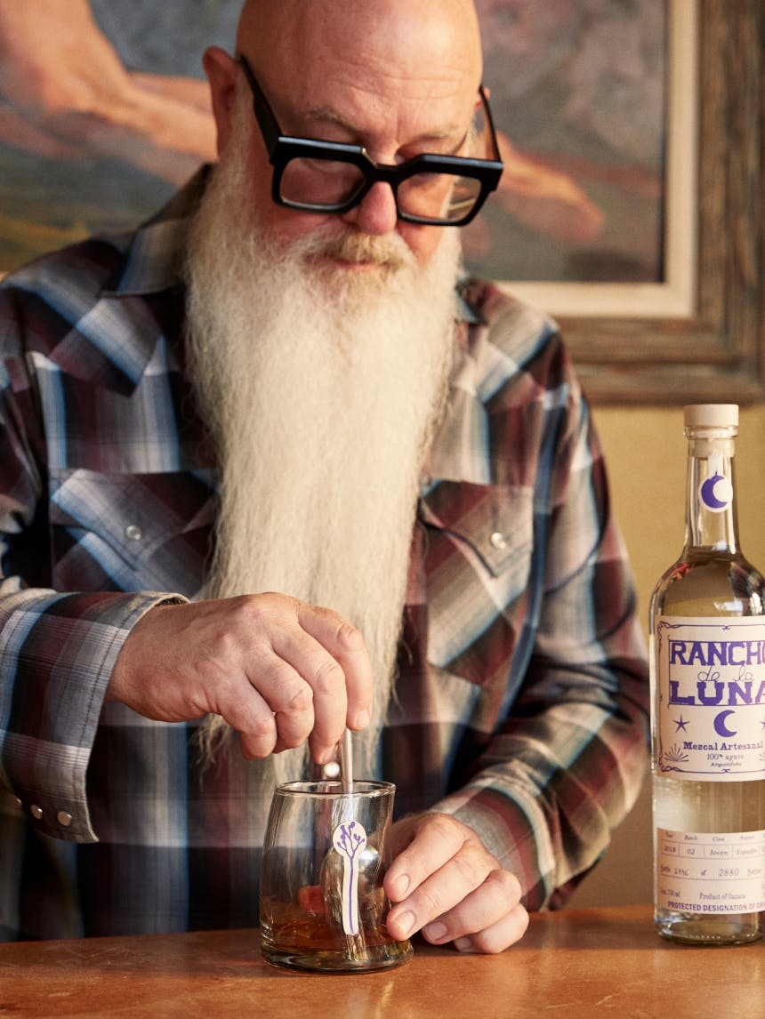 man with long beard muddling fruit in a glass next to a bottle of rancho de la luna mezcal