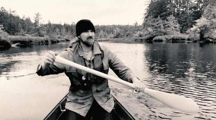 black and white image of man with a black beanie in a canoe holding a wooden oar in a boat on a river lined by pine trees
