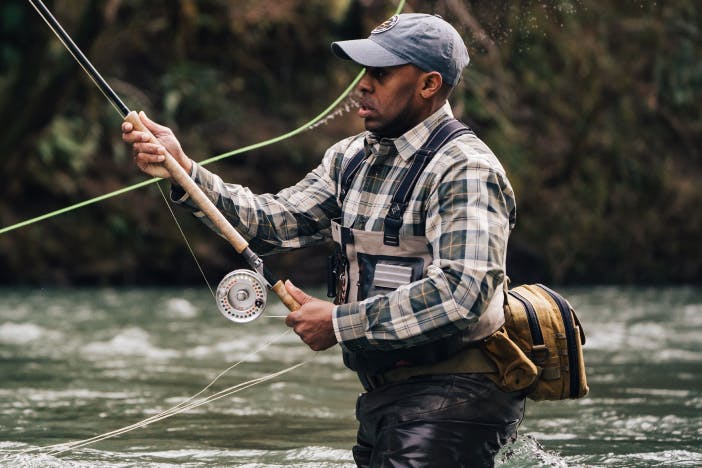 man wearing a green and white plaid shirt casting a fly fishing rod in knee high water