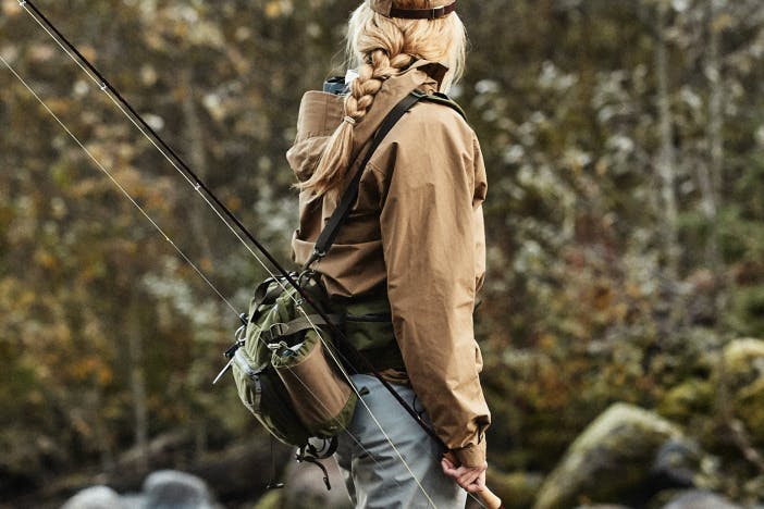 woman in a tan coat holding a fly fishing rod with a cross body filson pack on