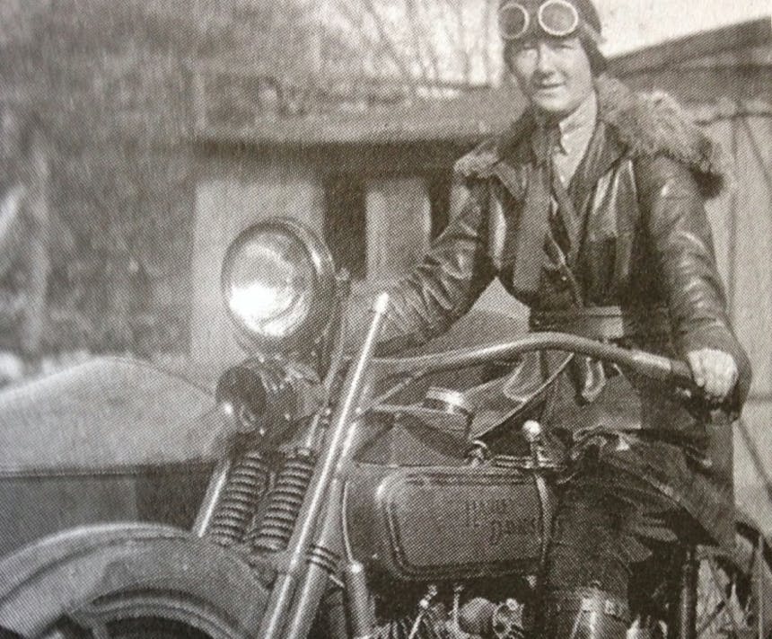 old black and white photo of a woman in antiquated motorcycle gear riding a vintage harley davidson