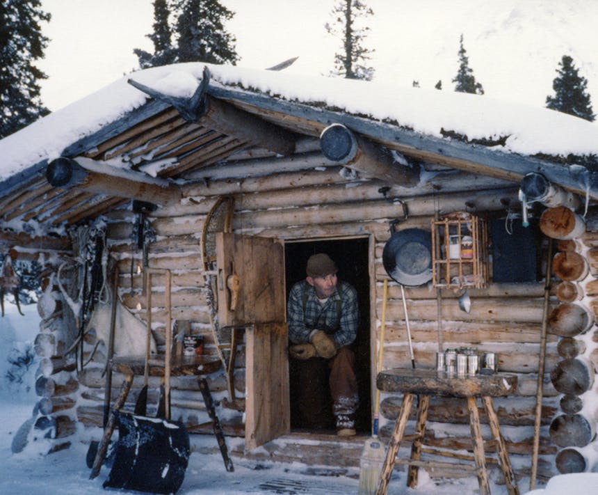 man in blue and white plaid shirt exiting a small door in a wood cabin with various cooking and hunting supplies outside on wooden tables and hanging