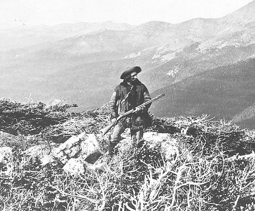 old black and white image of man in pancho and sombrero standing at a rocky outcropping with mountains in the distance