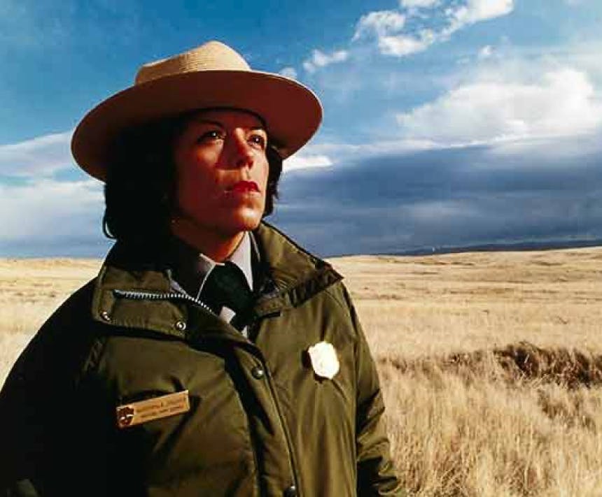 woman with white hat and green park ranger coat standing in a field of brown grass with cloudy skies on horizon