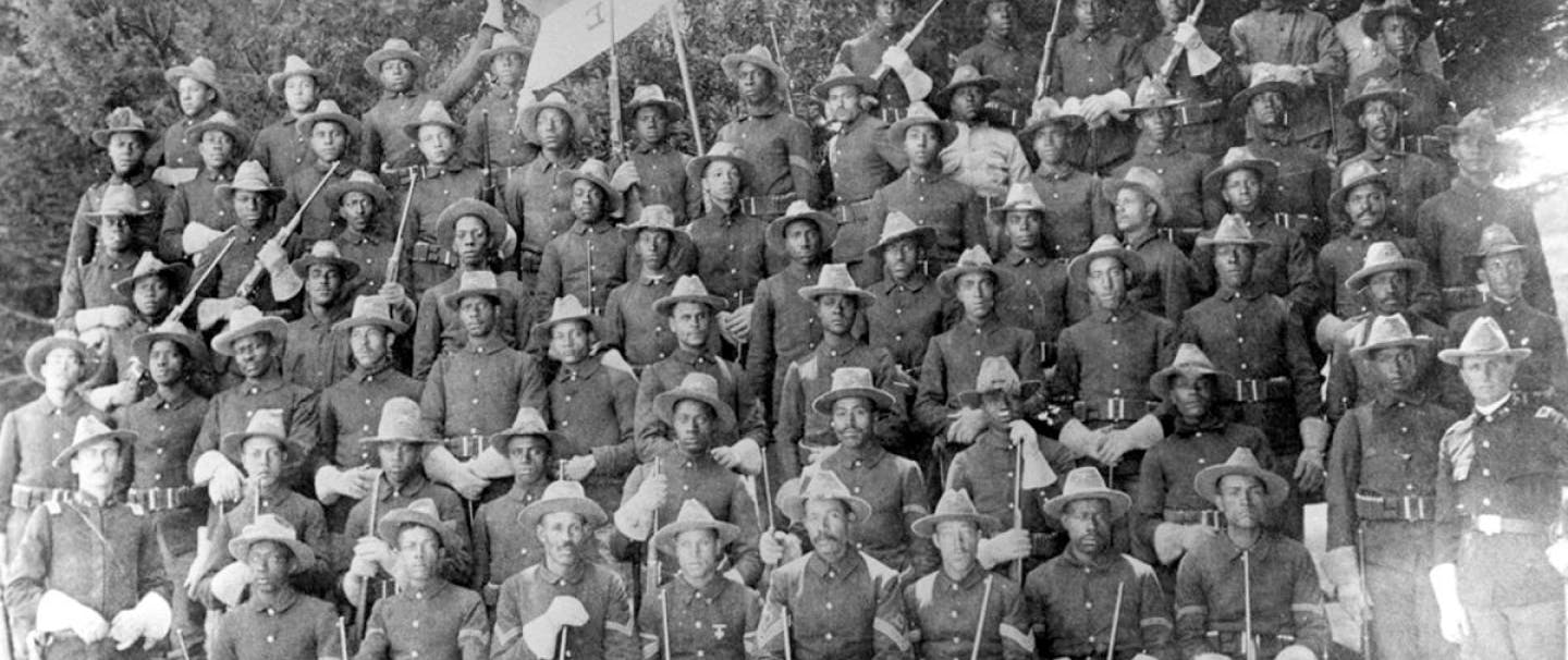 old black and white image of large group of people in dark uniforms with rifles and hats standing posing for a group picture
