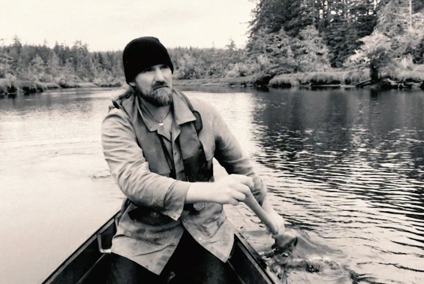 black and white image of a man in a tin cloth jacket paddling an oar while sitting in a boat on a river lined with pine trees