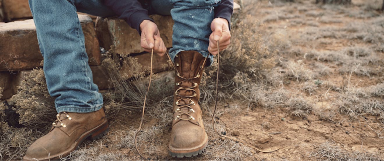 hands tying laces of brown leather boot on left foot while person sits on a set of stacked rocks