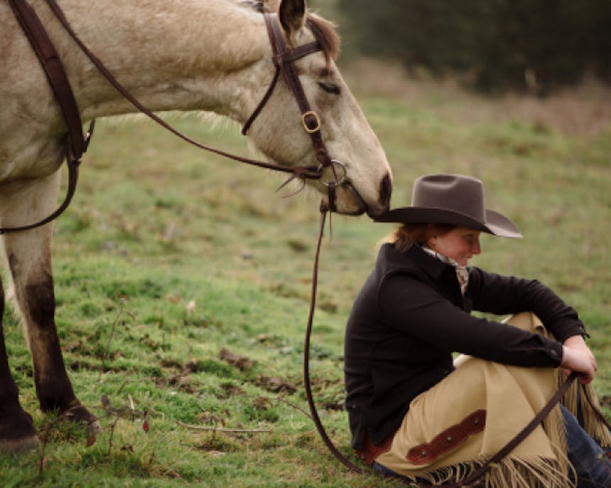 woman sitting by a white horse with tan chaps on and a gray hat