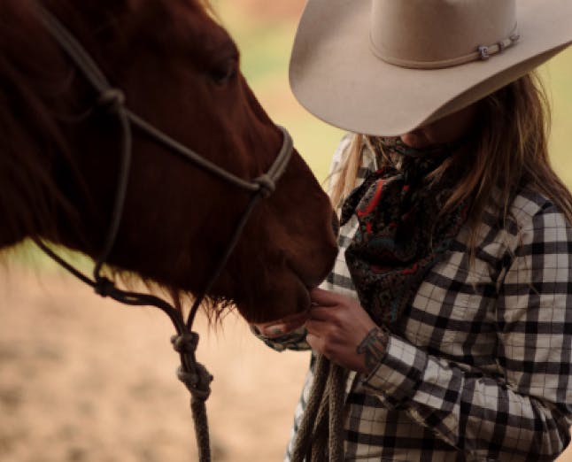woman in black and white plaid shirt and white hat feeding a horse