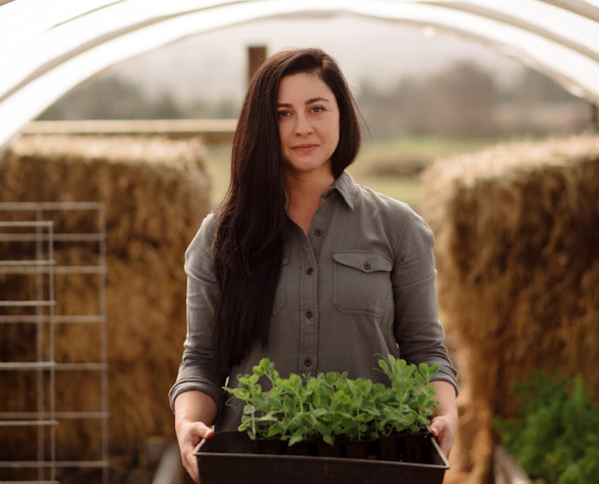 woman with long dark brown hair wearing a gray shirt holding a planter box with green herbs