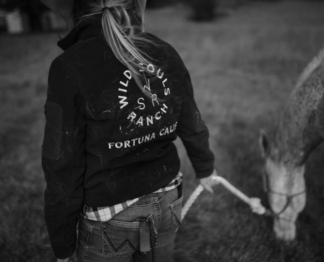 woman in a jacket with wild souls logo on the back leading a horse by it's reigns