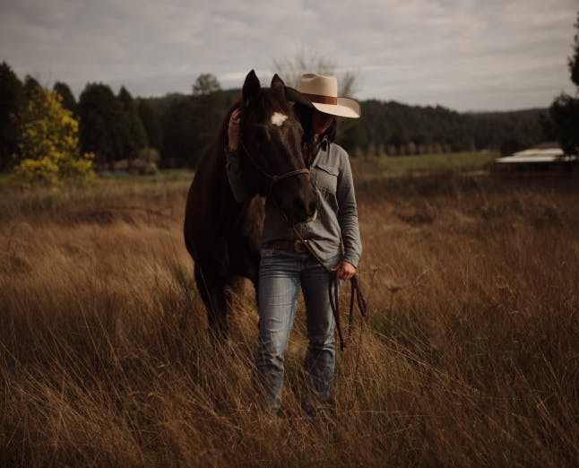 woman in white cowboy hat and gray jacket holding a horse around its neck standing in a grassy clearing