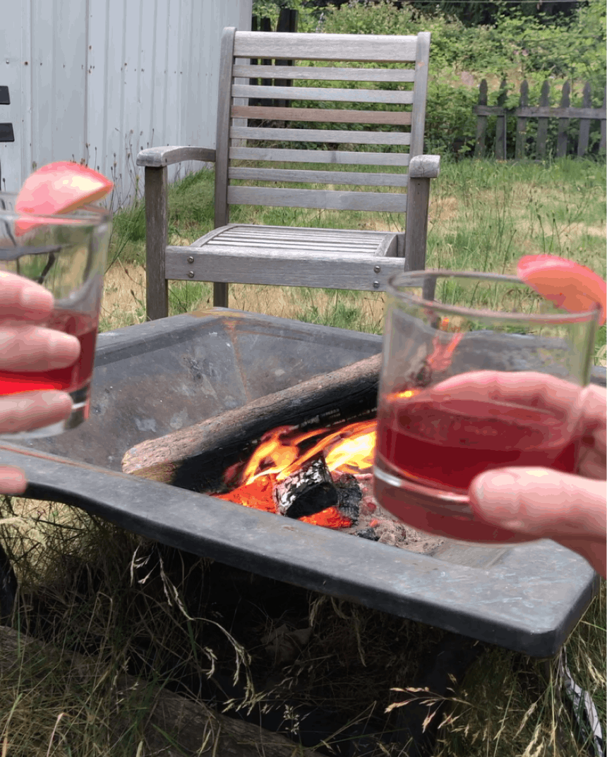 hands holding red cocktail in a rocks glass next to a firepit in a grassy lawn on a sunny day