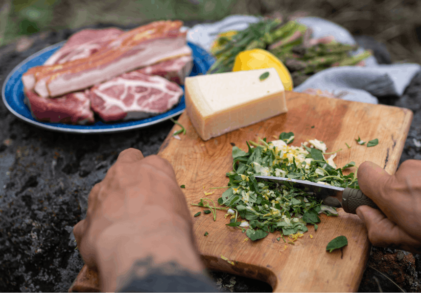 hand chopping herbs and lemon zest on a wooden cutting board next to a plate of pork shoulder, cheese, and foraged asparagus