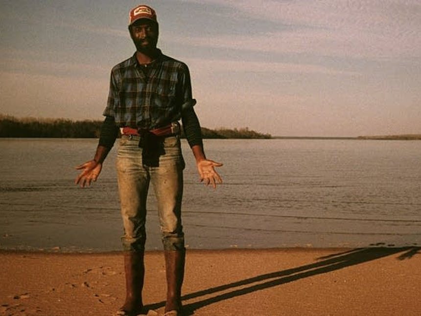 man in blue plaid shirt, waterproof boots standing on a beach at a lake