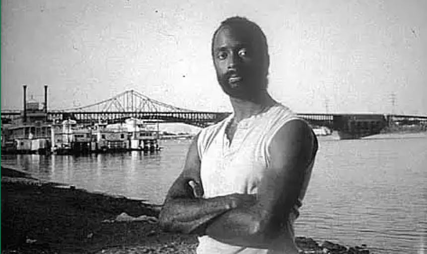 eddy harris standing on a beach in a tank top with an iron bridge and ships in the background