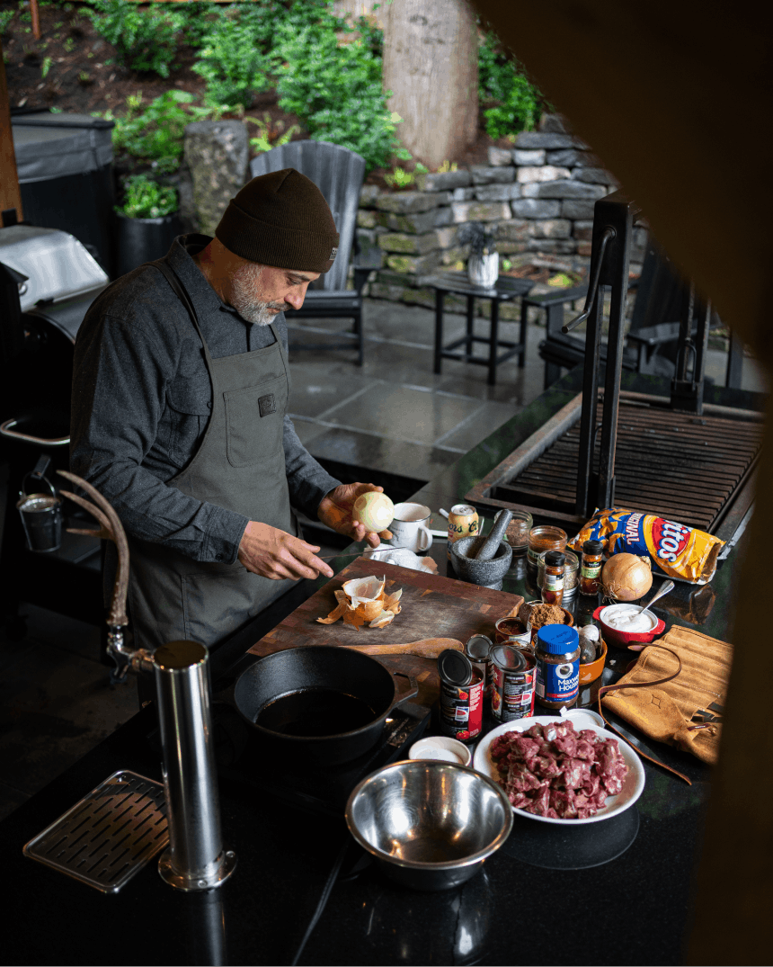 man in beanie and overalls holding an onion by his setup for cooking venison chili in a kitchen