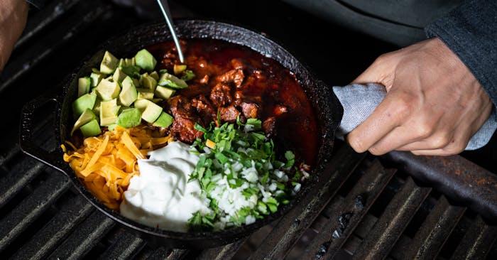 venison chili, cheese, avocado, sour cream, onion and herbs in a cast iron skillet on a grill grate