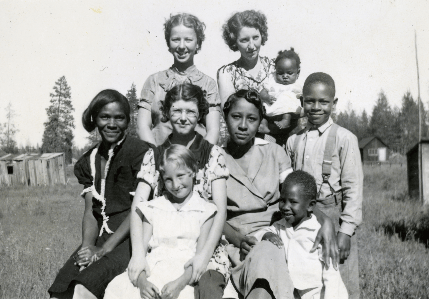 black and white image of a group of women and children standing in a field with a number of small structures spaced far apart from one another