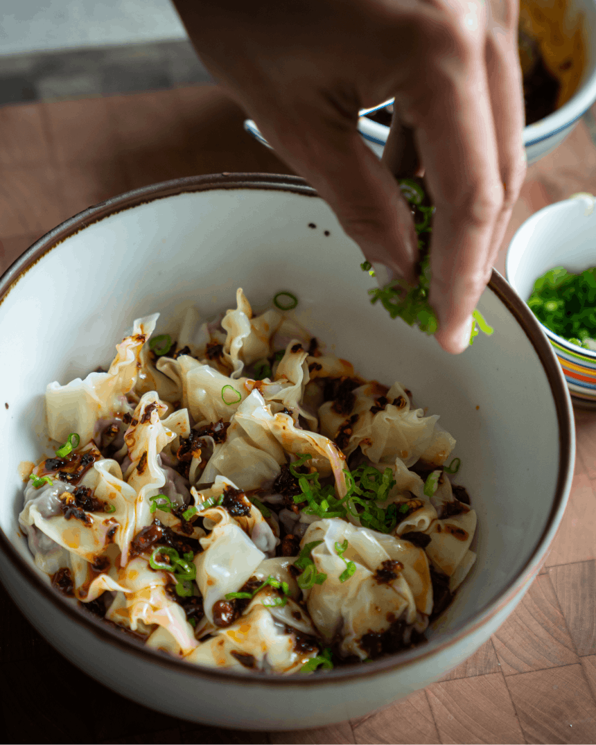 hands sprinkling scallions over wantons in a bowl
